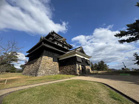 An historic Japanese castle. The sky is blue and the is a curved path leading towards the castle. It is wooden with a solid stone lower floor / foundation. It is white with black layered roofing.