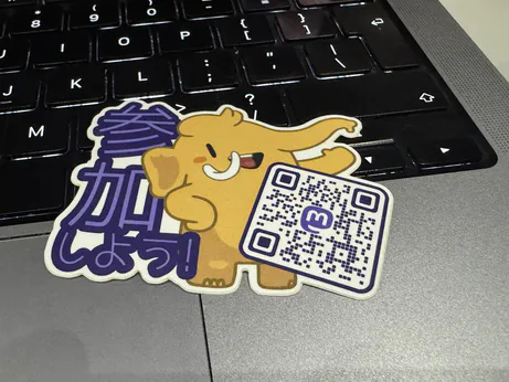 A sticker laying on the keyboard and trackpad of a MacBook laptop. The Mastodon mascot is in the centre, with a QR code that leads to joinmastodon.org to it's right, and some Japanese text 参加しよう! "let's participate!" in purple standing behind it on the left