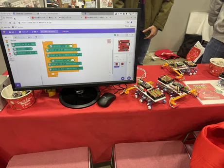 A screen showing a Scratch-like programming environment with Japanese text (smalruby) and some ESP32-based wheeled robots on a table.